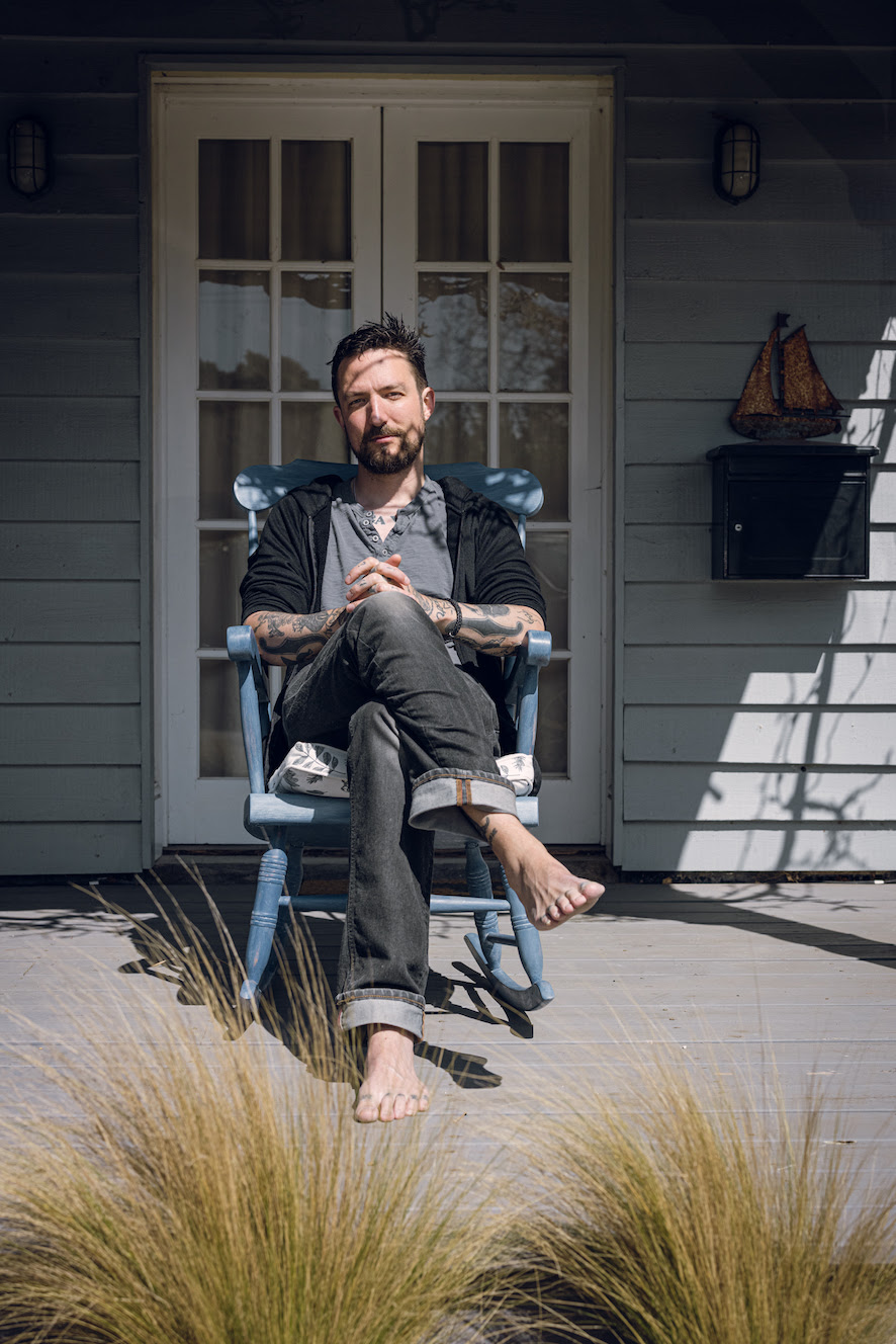 Frank Turner sitting in a blue wooden chair with no shoes or socks on