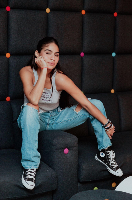 Jessie Reyez in jeans, vest top and converse sitting on the arm of a chair