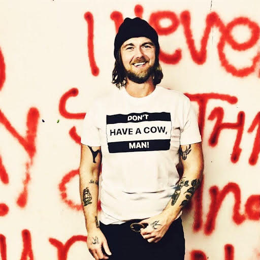 Charlie clark in front of a wall of graffiti with a white t-shirt saying Don't have a cow man on it