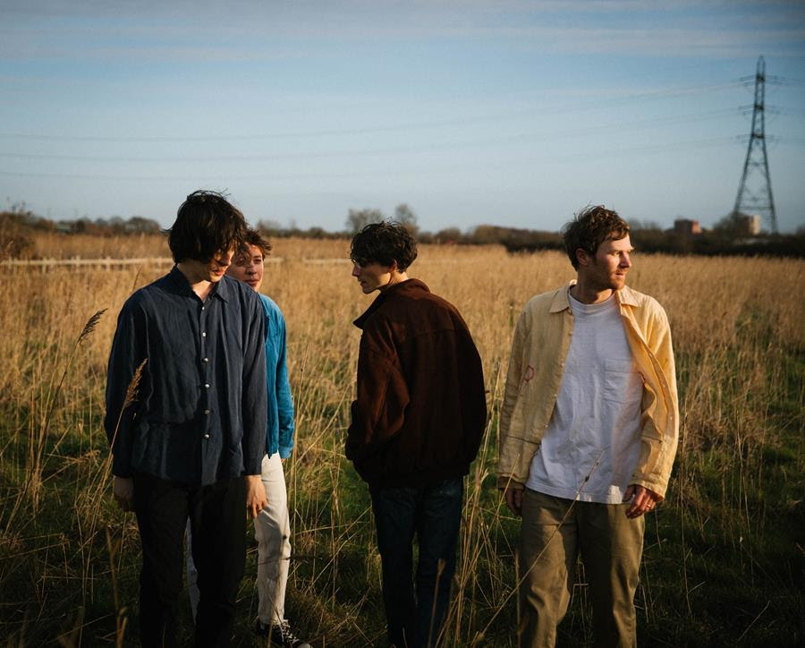4 members of Gengahr band standing in a field. Blue skies overhead and an electricity pylon in the background.