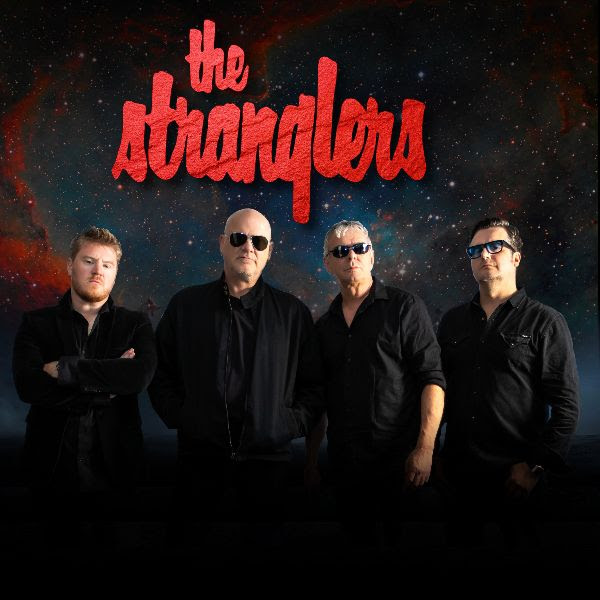 4 members of The Stranglers all in black shirts stand in front of a background of the galaxy