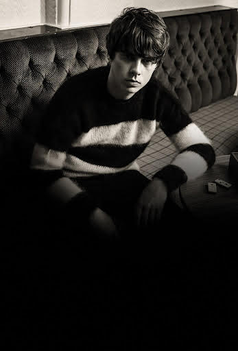 Jake bugg sitting on a long sofa with a striped jumper on