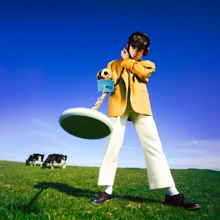 Declan McKenna album cover with a man using a metal detector in a field with 2 cows behind him