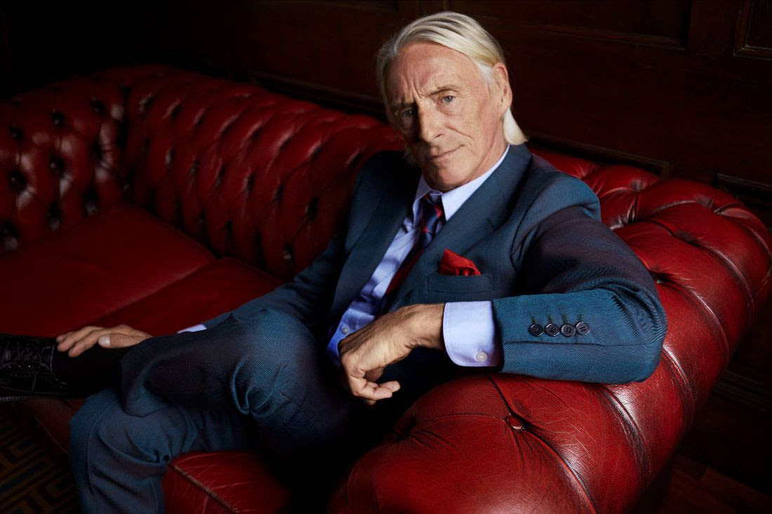 Paul Weller sat on a red leather sofa with a blue suit on