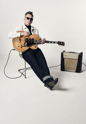 Richard hawley sat in a chair holding a guitar and an amp set up next to him