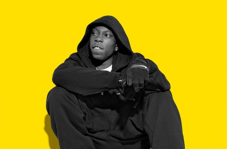 Dizzee Rascal sitting against a yellow background in a dark hoodie
