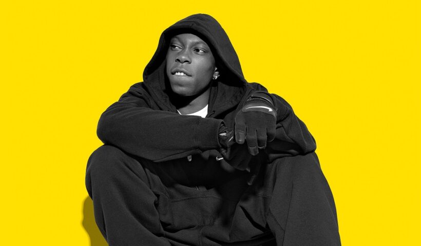 Dizzee Rascal sitting against a yellow background in a dark hoodie