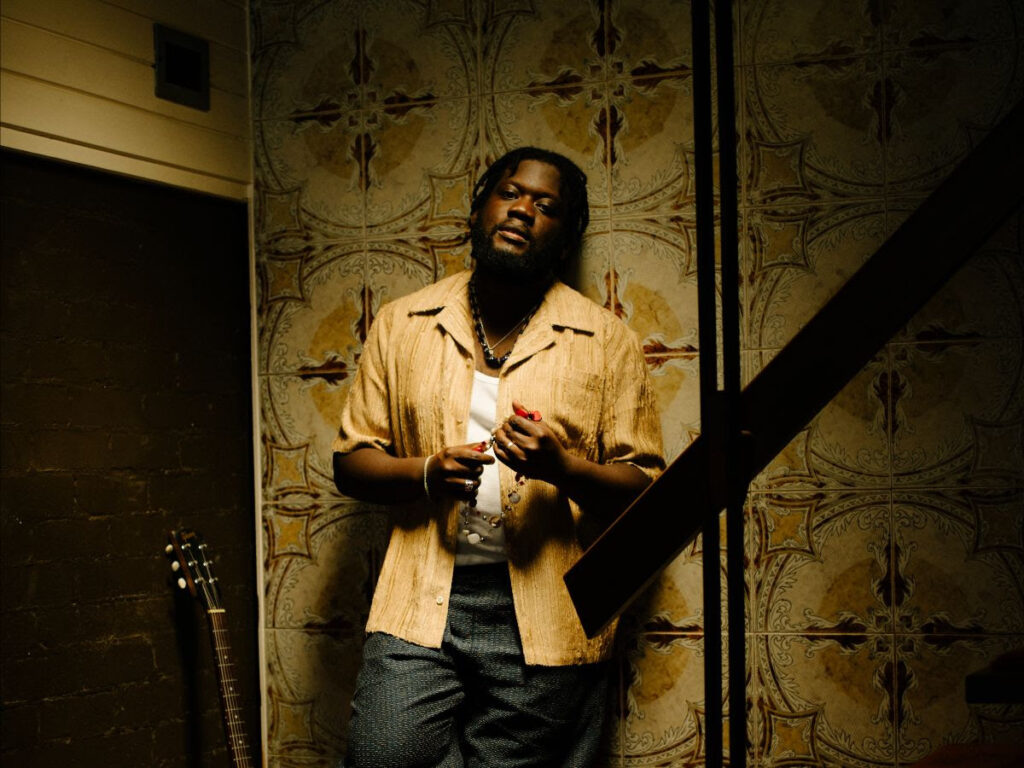 Michael Kiwanuka stood against a wall with his guitar leaning on an opposite wall