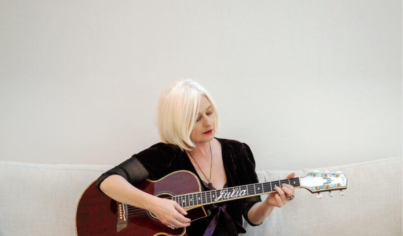 Julie Fordham sitting on a white sofa playing her guitar in a white room