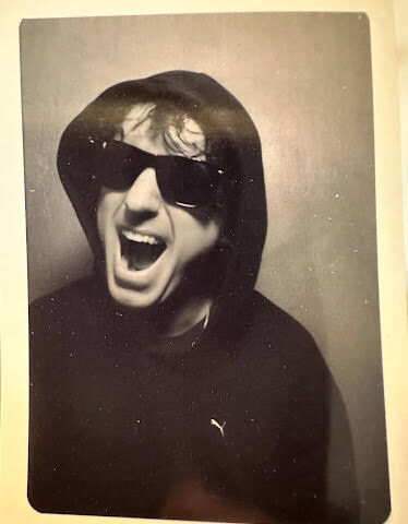 Johnny Lloyd in a hoody and sunglasses with his mouth wide open in a photo booth