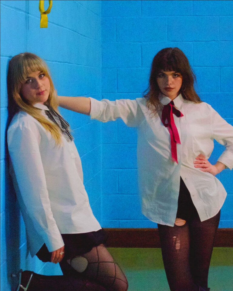 Both female members of the pill leaning against a wall dress in school uniforms
