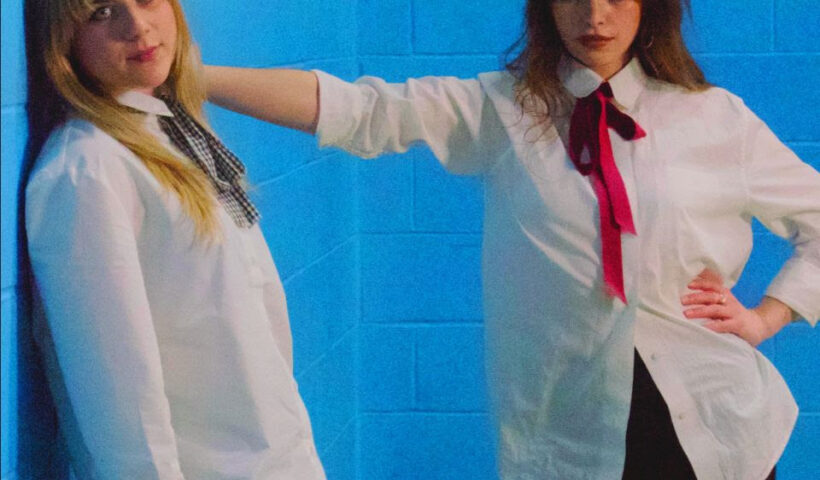Both female members of the pill leaning against a wall dress in school uniforms