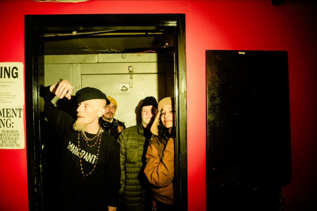 Stereo MCs in a doorway looking out. Lead singer is leaning on the frame of the doorway