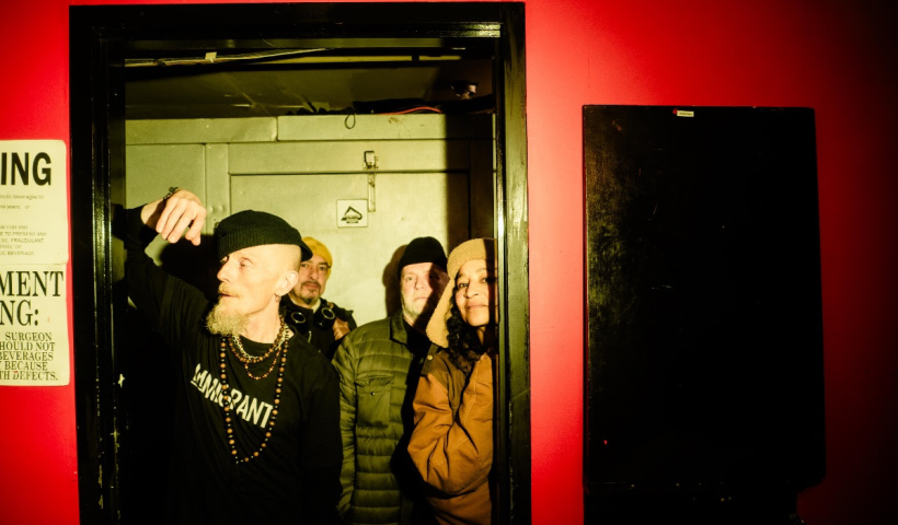 Stereo MCs in a doorway looking out. Lead singer is leaning on the frame of the doorway