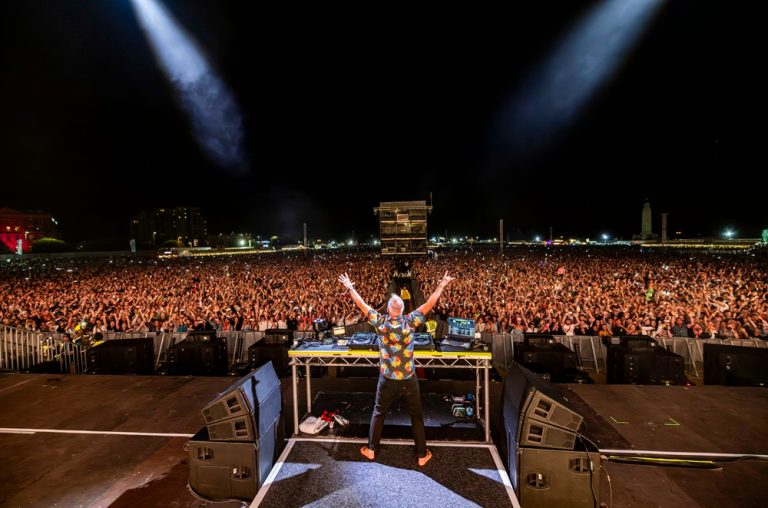 Photo of fatboy slim from behind with his arms raised in front of thousands of people