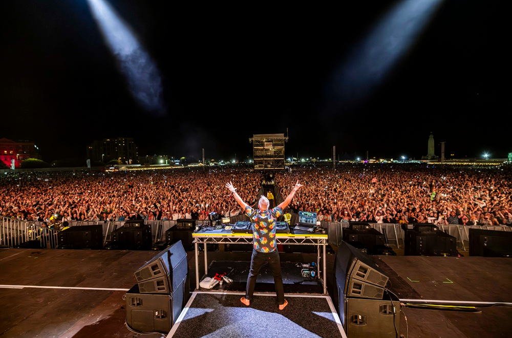 Photo of fatboy slim from behind with his arms raised in front of thousands of people
