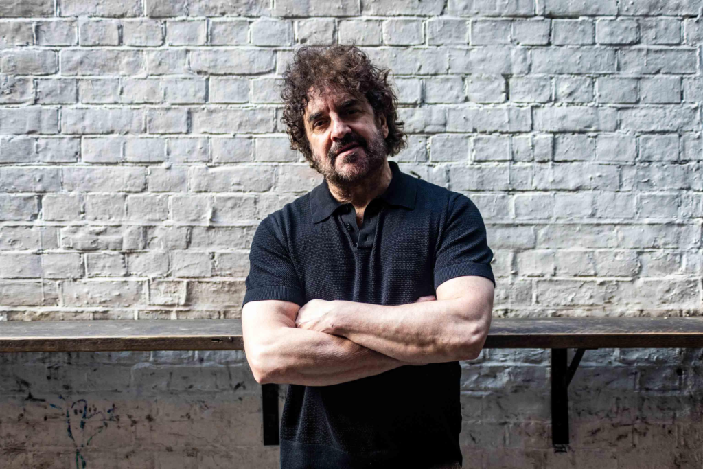 Ian Prowse stood with his arms folded with a white brick wall behind him