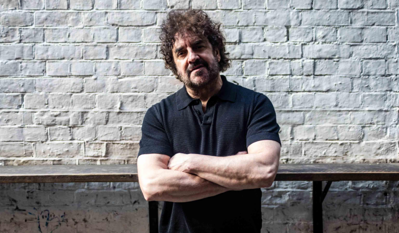 Ian Prowse stood with his arms folded with a white brick wall behind him